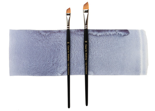 Two angle watercolor brushes with black handles and orange-brown tips, standing upright on a rectangular sheet of textured paper showing a periwinkle-blue wash, against a white background.