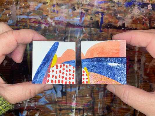 Two hands hold two small square abstract paintings side-by-side. The paintings feature bold brushstrokes of blue, orange, red, yellow, and white.