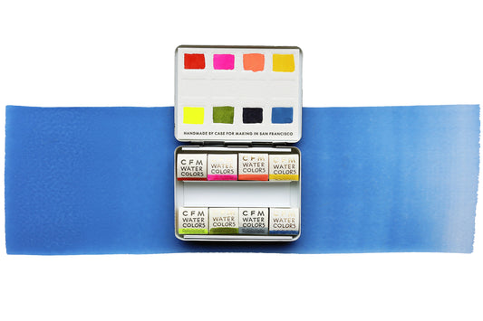 A handmade watercolor palette featuring Case for Making Colors: French Vermilion, CfM Hot Pink, CfM Coral, CfM Oxalis, Fluorescent Yellow 2, CfM Olive, Synthetic Indigo, CfM Shadow Blue open on a blue painted swipe on a white background
