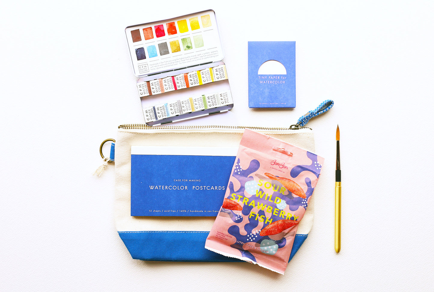 Photo of the Summer Bundle. Top down view starting with the 14 color combo small palette in the top left, next to the CfM Tiny paper. Below that, the CfM Postcards with the bag of Sour gummies are on top of the canvas bag with a gold travel brush to the right.