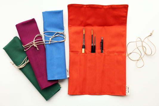 A set of fabric art supply rolls in multiple colors, including orange, blue, purple, and green, shown on a white background. The orange roll is open, displaying pockets holding paintbrushes and a pencil, with a beige leather cord tied around it.