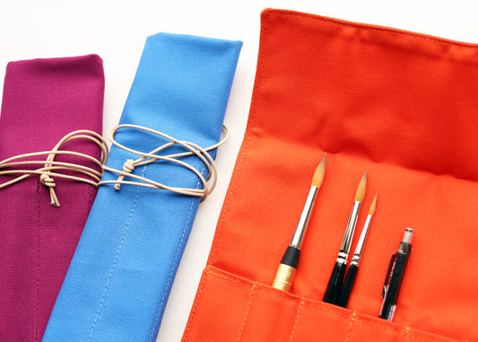 An orange fabric art supply roll with several pockets holding paintbrushes and a pen, shown next to purple and blue rolls tied with cord on a white background.