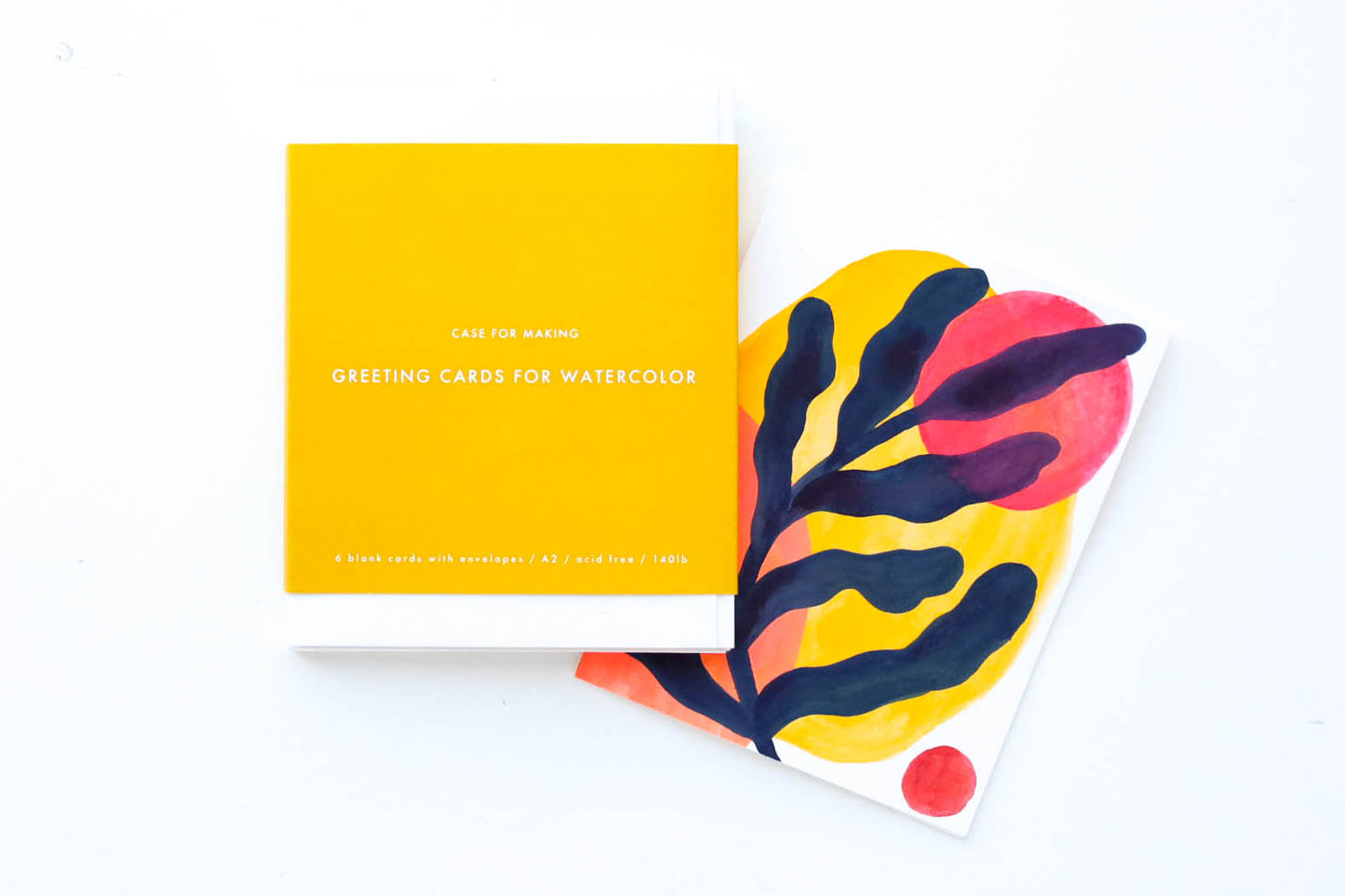 Case for Making Greeting Cards for Watercolor Pack. Six stacked white cold press watercolor folded cards with accompanying white envelopes. Packaged and wrapped in a golden yellow band of paper with the branding printed in white. Also shown is a sample card with a painting on the cover.