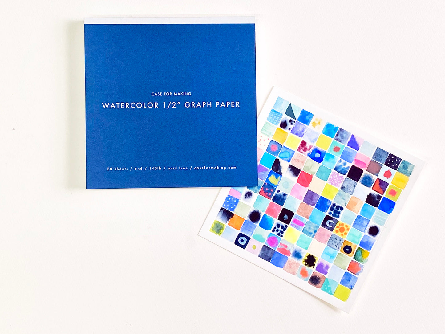 The closed, top-bound 6" x 6" Watercolor 1/2" Graph Paper pad. Showing the dark blue cover with white lettering. Also shown is a painted sample sheet of the 1/2" Graph Paper.
