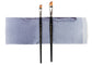 Two angle watercolor brushes with black handles and orange-brown tips, standing upright on a rectangular sheet of textured paper showing a periwinkle-blue wash, against a white background.
