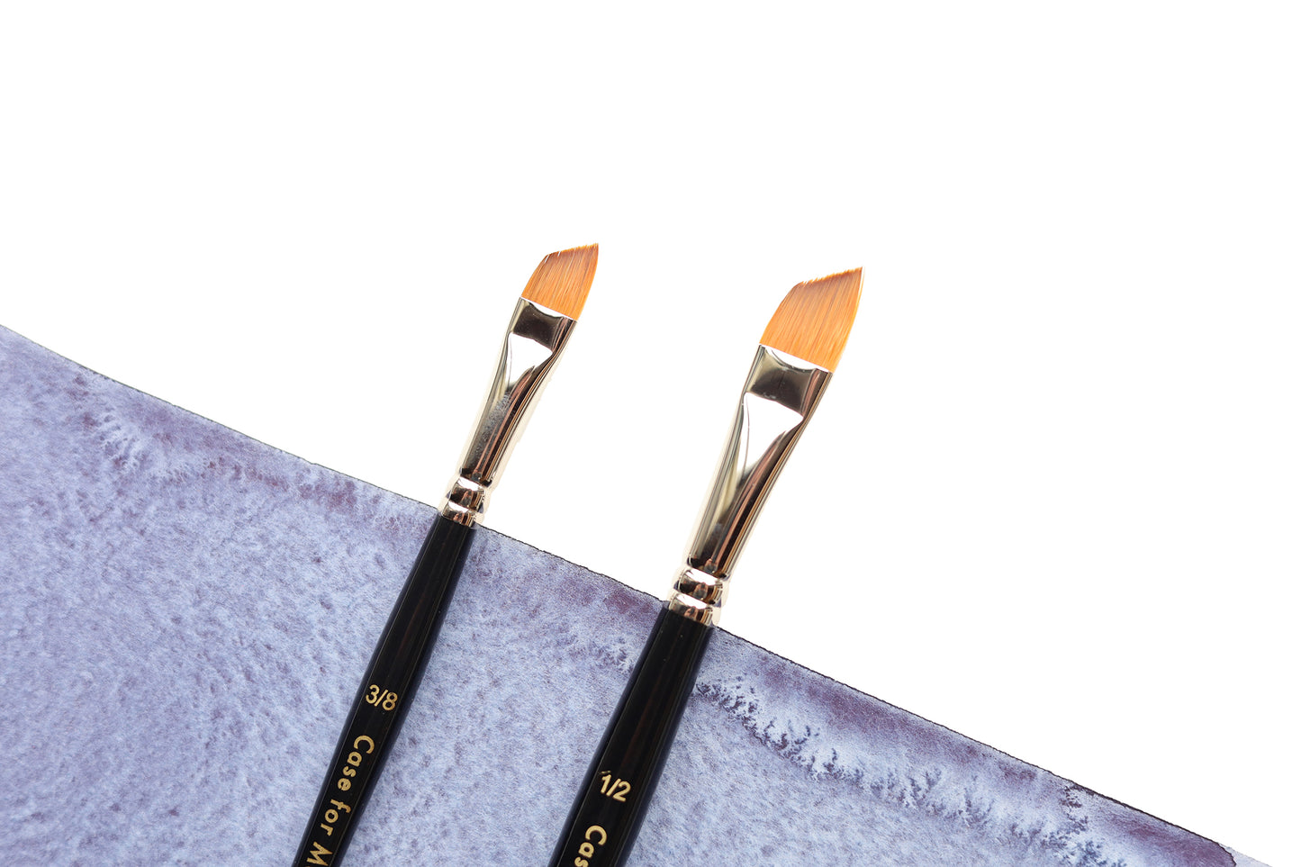 Two angle watercolor brushes with black handles and gold ferrules resting on a textured light indigo watercolor wash against a white background.