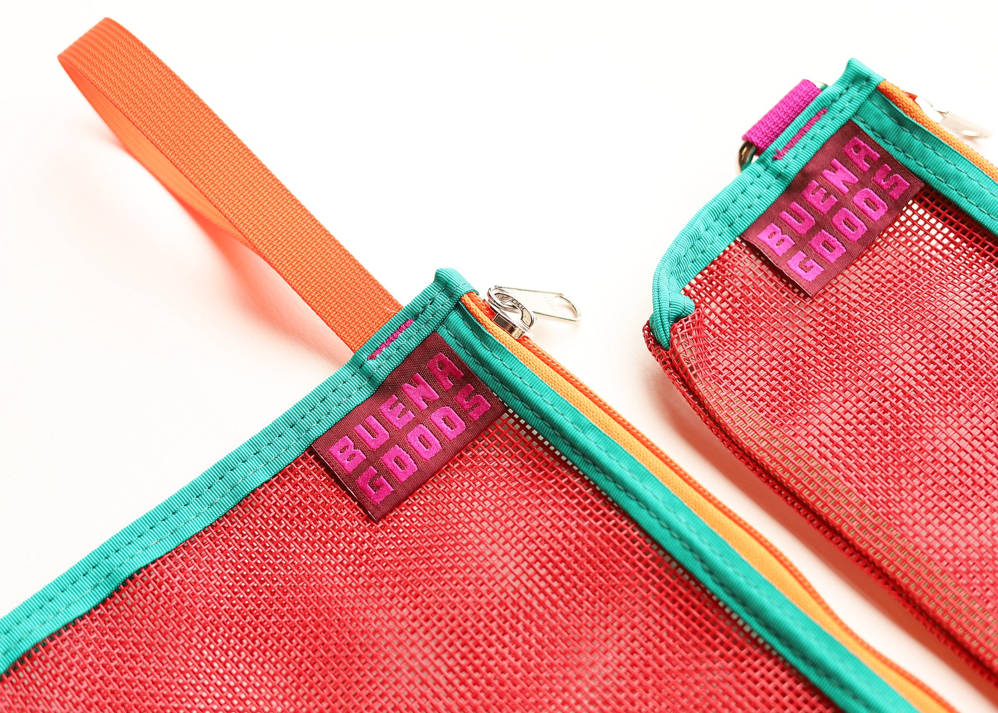 Close-up of two mesh zip pouches in bright coral red with teal trim and orange webbing handles, both showing a purple Buena Goods brand tag.