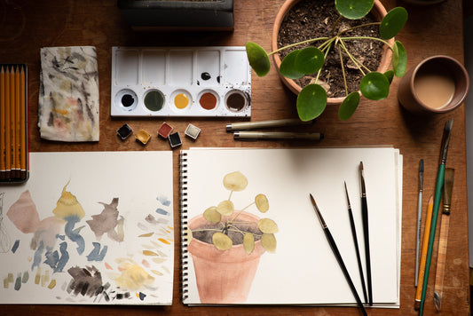 A styled flat-lay of art supplies on a wooden table, including two sketchbooks with watercolor paintings of a potted plant, paintbrushes, a watercolor palette, pencils, and a small potted plant.