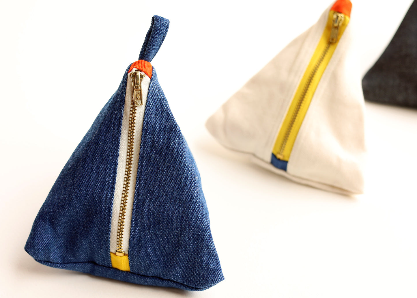 Two triangular fabric pouches with zippered closures on a white background. The front pouch is made of blue denim with a gold zipper, white trim, and a small orange tab.