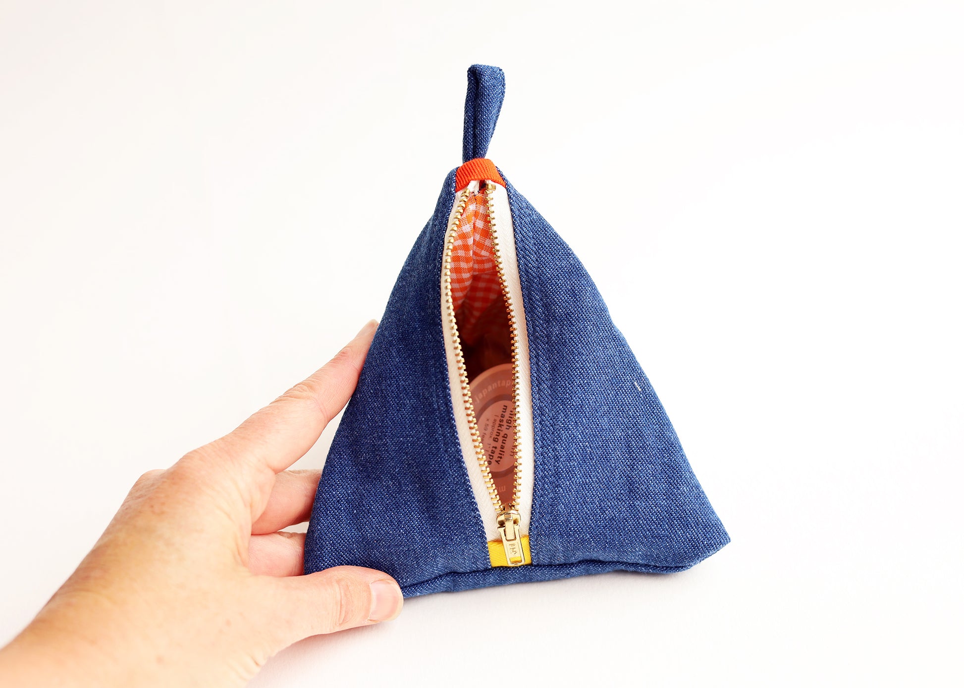 A hand holding a small triangular blue fabric pouch with a top loop and an open gold zipper showing an orange patterned lining, against a plain white background.