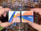 Two hands hold two small square abstract paintings side-by-side. The paintings feature bold brushstrokes of blue, orange, red, yellow, and white.