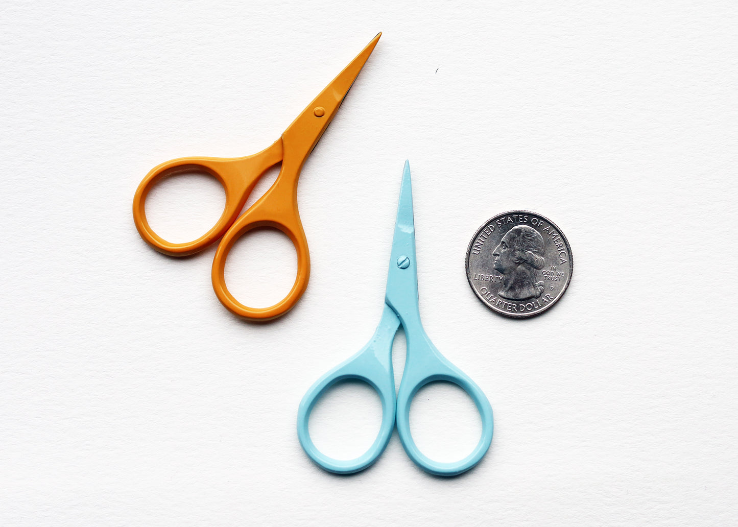 Two small scissors on a white textured background, one with orange handles and one with light blue handles, with a US quarter coin placed next to the blue scissors for scale.