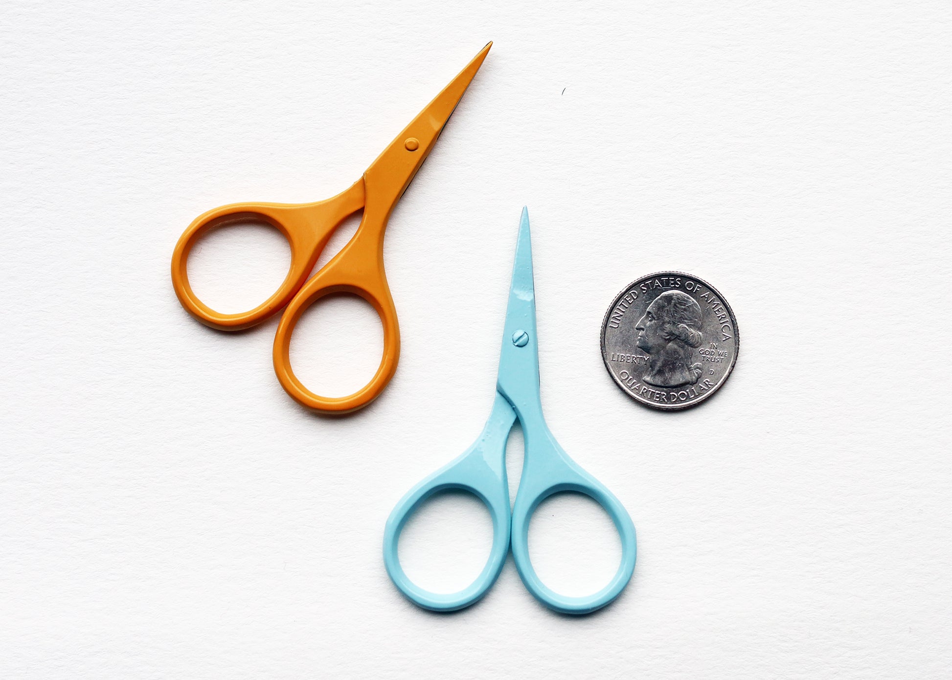 Two small scissors on a white textured background, one with orange handles and one with light blue handles, with a US quarter coin placed next to the blue scissors for scale.