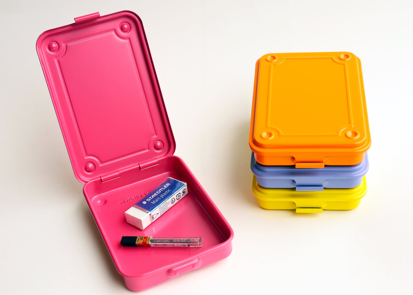 A stack of four rectangular stamped metal storage in bright pink, orange, lilac, and yellow, photographed on a clean white background. One box is open showing an eraser and pack of pencil lead.