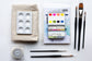 An image of watercolor supplies on a white background including 5 brushes, a watercolor palette, paper, mixing palette, tape and dish cloth.