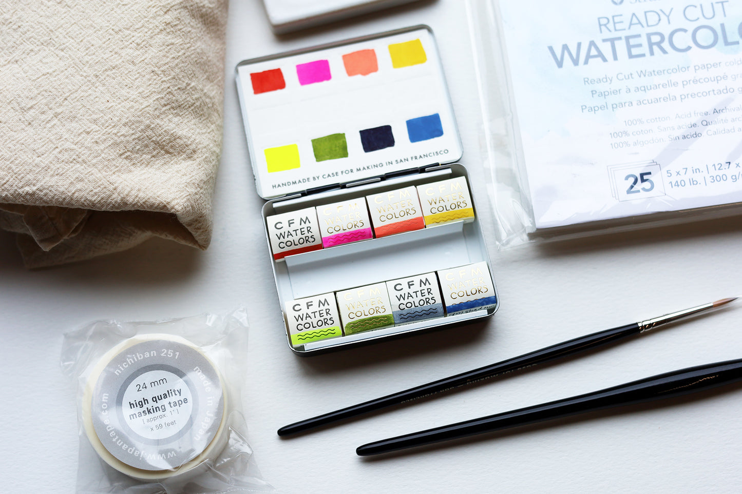 An image of watercolor supplies on a white background including 5 brushes, a watercolor palette, paper, mixing palette, tape and dish cloth.