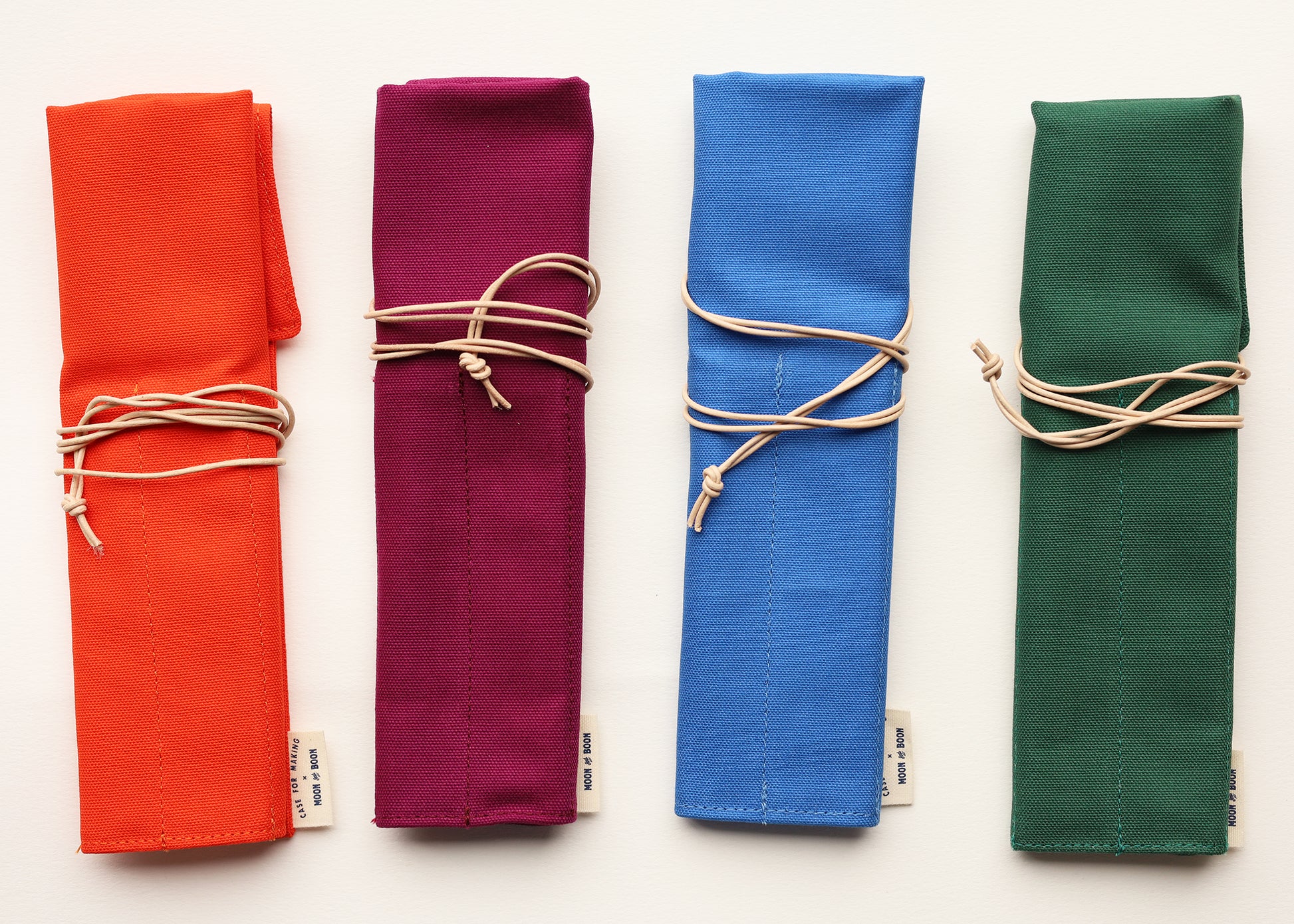 Four rolled fabric napkins in orange, raspberry, blue, and green, each tied with a beige leather string and shown on a white background.