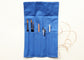 A blue fabric art supply roll-up pouch with multiple pockets holding paintbrushes, a pencil, markers, and a ruler, shown on a white background with a beige cord attached.