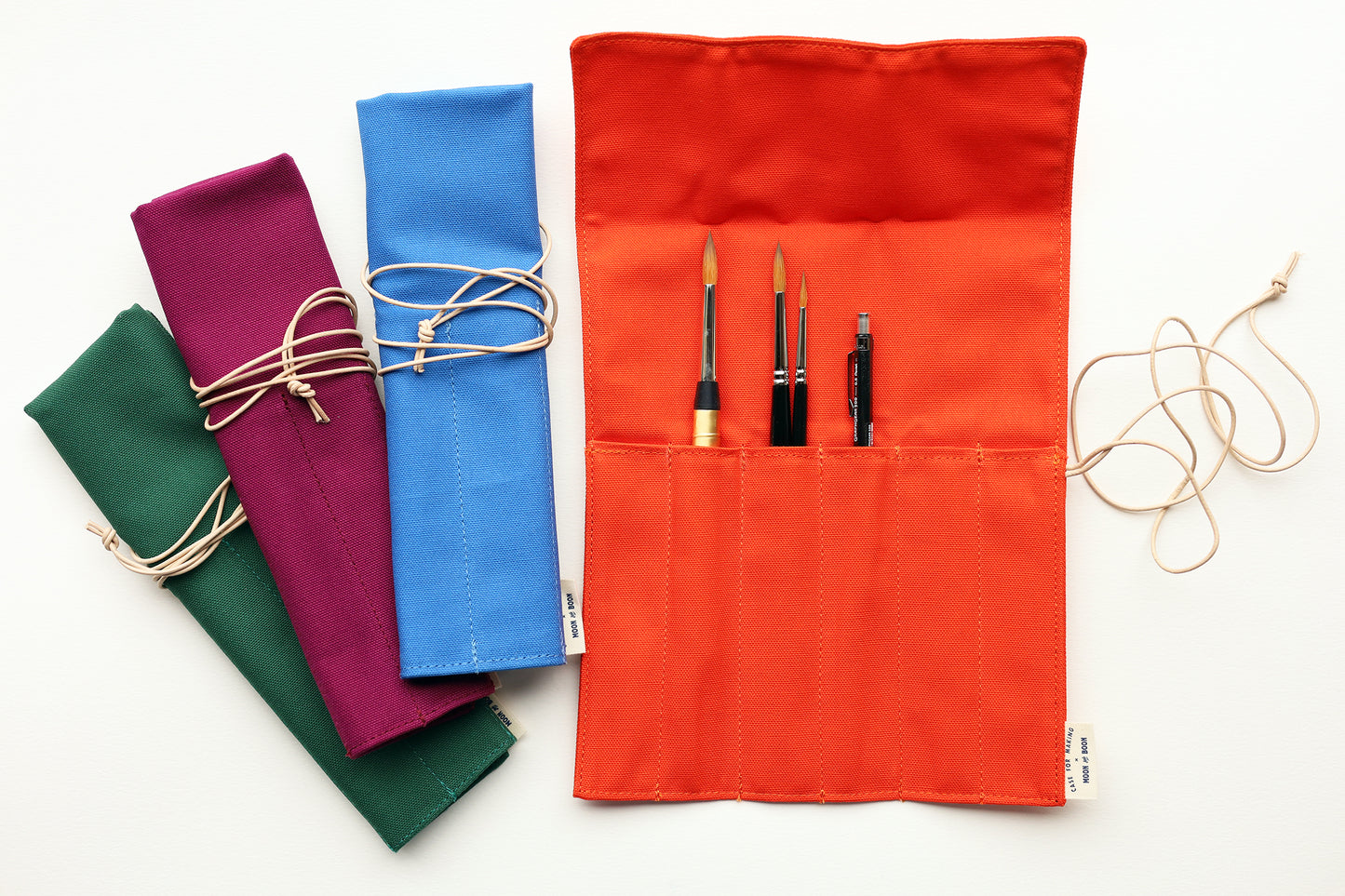 A set of fabric art supply rolls in multiple colors, including orange, blue, purple, and green, shown on a white background. The orange roll is open, displaying pockets holding paintbrushes and a pencil, with a beige  leather cord tied around it.