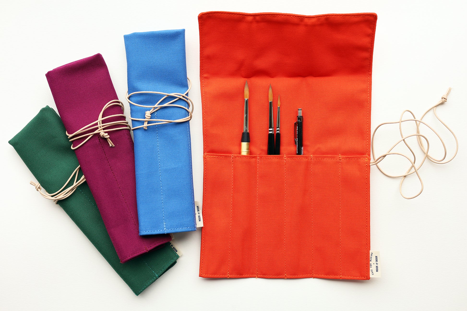 A set of fabric art supply rolls in multiple colors, including orange, blue, purple, and green, shown on a white background. The orange roll is open, displaying pockets holding paintbrushes and a pencil, with a beige  leather cord tied around it.