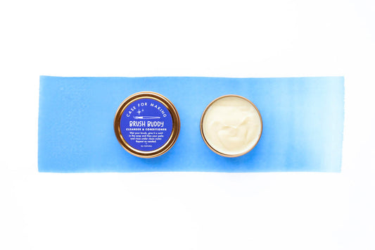 One closed Brush Buddy, showing the lid with the Ultramarine Blue label with white lettering, beside one opened Brush Buddy showing the light cream colored cleanser.